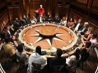 The Traitors season 2 cast sits around the roundtable listening to host Alan Cumming during season 1, episode 2