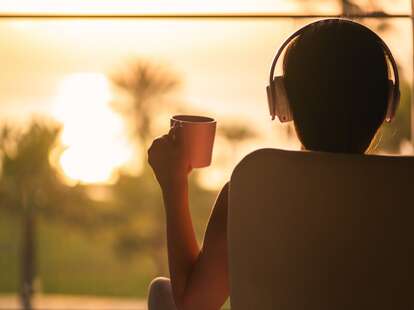 Rear view of woman with headphones and cup of coffee sitting on chair near window and looking on palm trees