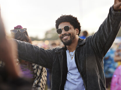 Happy young man wearing sunglasses dancing with arms raised at music festival during sunset.