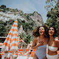 Two women look delighted to be holidaying together in Italy. Views of Positano are visible behind them. They are surrounded by striped beach umbrellas and sun loungers. They wear stylish swimwear.