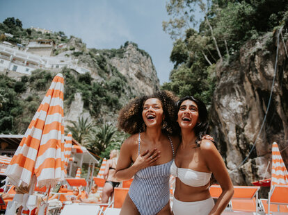 Two women look delighted to be holidaying together in Italy. Views of Positano are visible behind them. They are surrounded by striped beach umbrellas and sun loungers. They wear stylish swimwear.