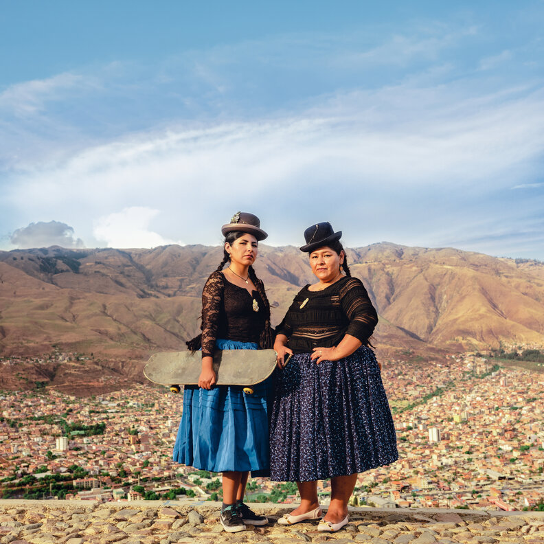 two women in traditional indigenous skirts. One holds a skateboard.