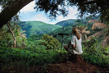 a woman with a telescope in a green forested and hilly landscape