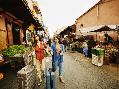 Wide shot of female friends walking with luggage through the souks of Marrakech while on vacation.