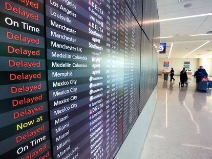 Passengers walk past a flight status board in Terminal C at Orlando International Airport that shows many delays.