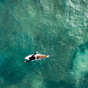An aerial view of a woman on a surfboard paddling out to the waves while surfing in a tropical setting. 