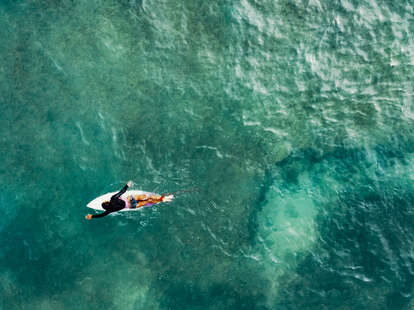 An aerial view of a woman on a surfboard paddling out to the waves while surfing in a tropical setting. 