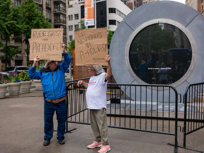 Two men hold signs in front of a video “Portal” art installation connecting New York City and Dublin, Ireland via a 24/7 video livestream which sits temporarily shut down in the Flatiron Plaza on May 15, 2024 in New York City.