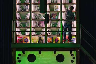 a man standing in front of an elevated wall filled with vinyl records