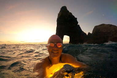 woman swimming in goggles while sun sets behind her and rock formation