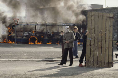 Young people on the streets of Belfast in 1972, standing in front of a burning bus. One young man is wearing a face covering and holding a rock.