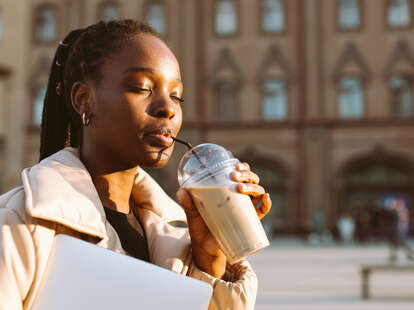 Young woman drinking an iced latte from disposable cup standing in town square during daytime