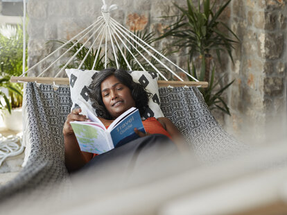 Smiling mature woman reading book while lying in hammock on vacation.