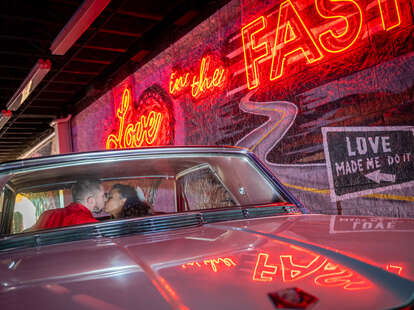 a couple kissing in a cadillac in front of a neon sign that reads "Love in the Fast Lane"