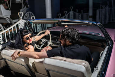 a man and a woman wearing black, in a pink cadillac. the woman is looking towards the camera