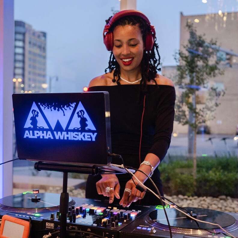 A woman behind DJ decks and a screen that says Alpha Whiskey