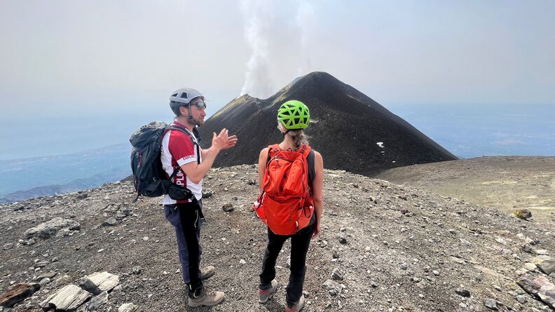 two people standing on mountain with a volcano smoking in the background