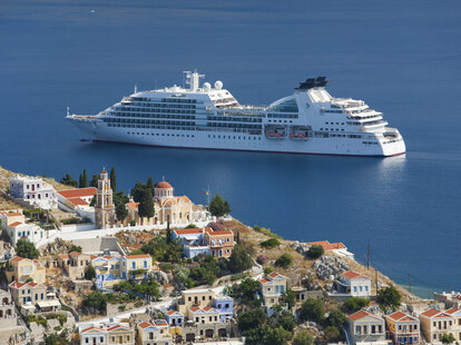 Hilltop houses and Greek Orthodox church dwarfed by cruise ship anchored offshore, Gialos (aka Yialos), Symi (aka Simi), Rhodes, Dodecanese Islands, South Aegean, Greece, Europe.