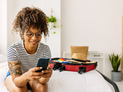 Happy young woman booking a hotel room while packing her summer suitcase for holidays