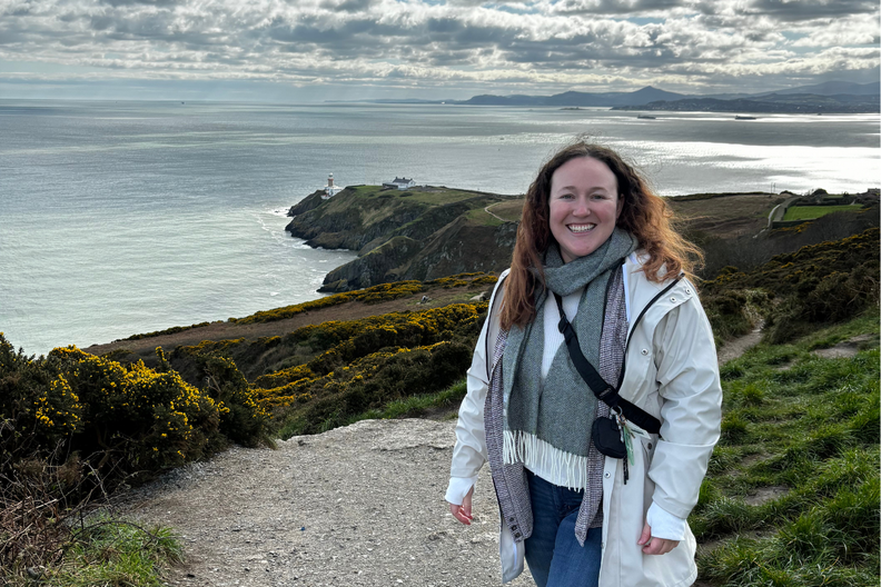 The writer in Howth, Ireland, standing in front of cliffs and the water during the day.