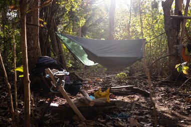 The makeshift survival camp, with hammocks hanging in the tree in the forest and debris on the floor.
