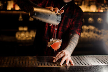 a bartender pouring a pink drink into a cocktail glass decorated with a paper plane