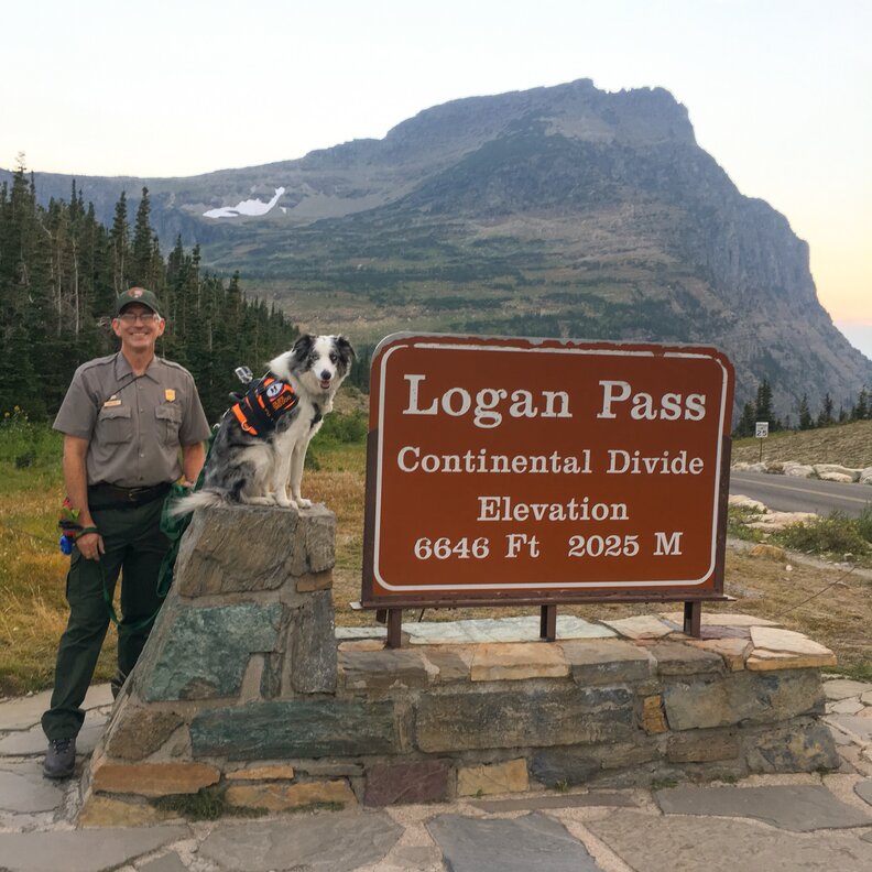a man and a dog at the Logan Pass Continental Divide sign at Glacier National Park