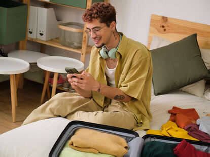 Young man is using a smartphone while packing a suitcase on the bed.