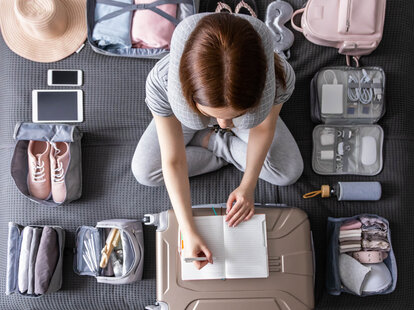 Smiling tourist woman packing suitcase to vacation writing paper list sitting on bed at home. Female getting ready to travel trip organization things storage in comfy cases bags use konmari method.