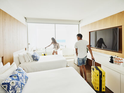 Wide shot of man and woman rolling luggage into hotel room at tropical resort on vacation.