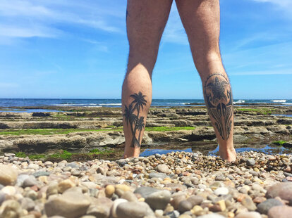 nude man with leg tattoos standing before the beach