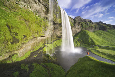 a small man in a red jacket standing next to Seljalandsfoss waterfall in iceland on a sunny day