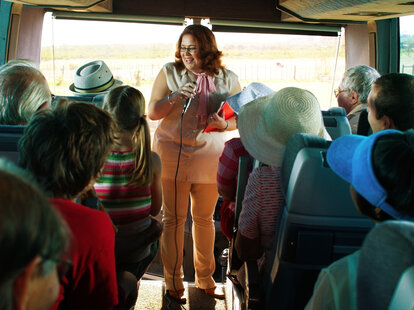 Tour guide holding microphone at front of coach bus full of tourists.