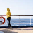 A cruise passenger in a yellow parka stands at the railing at the stern of a cruise ship and looks back at the blue open sea. Next to the person hangs an orange life preserver