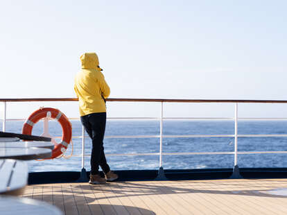 A cruise passenger in a yellow parka stands at the railing at the stern of a cruise ship and looks back at the blue open sea. Next to the person hangs an orange life preserver