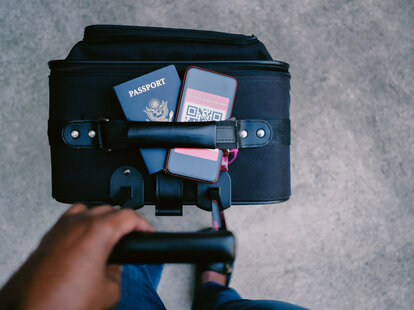 High angle view person standing with passport and COVID-19 digital vaccine on mobile phone tucked underneath luggage handle.