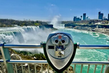 On a bright blue and sunny day, binoculars point towards a majestic waterfall. Mist sprays into the air in the distance as water pours over the side of the cliff and falls to the gorge below.