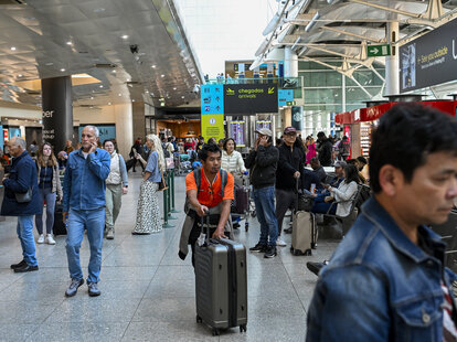 A person in a brightly colored shirt pushes a suitcase along a train station as the surrounding crowd moves around them.