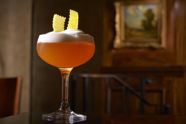 cocktail in a coupe glass with a lemon rind garnish on a dark wood table in front of an old landscape painting in the rosewood mansion in dallas