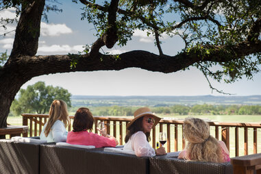 four women sit on couches overlooking a vineyard in texas wine country, under an oak tree
