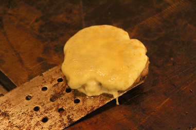 A spatula picks up PYT's provolone topped lasagna bun burger from the grill.
