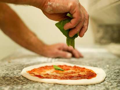 A hand drops fresh basil unto an uncooked pizza smeared with tomato sauce.